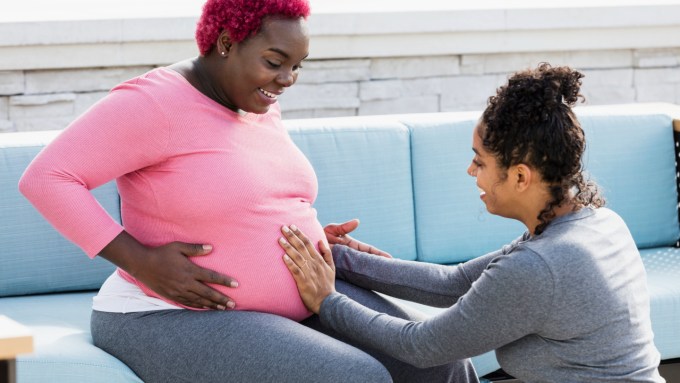 A doula is knelt in front of a pregnant person, who is sitting on a blue couch, and has their hand placed on their stomach.